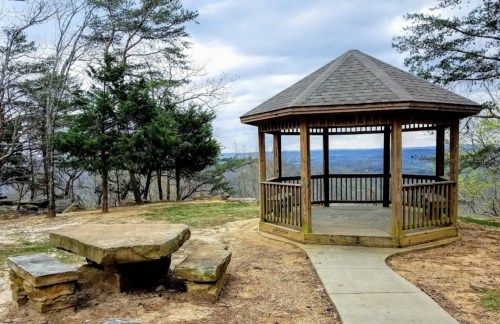 A wooden gazebo on a hillside with a stone table, surrounded by trees and a cloudy sky in the background.