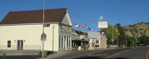 A small town street featuring a motel, shops, and flags from various countries, with mountains in the background.