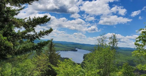 A scenic view of a lake surrounded by lush green trees and rolling hills under a bright blue sky with fluffy clouds.