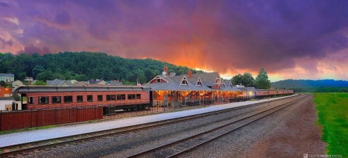 A scenic train station at sunset, with vibrant clouds and lush greenery along the tracks.