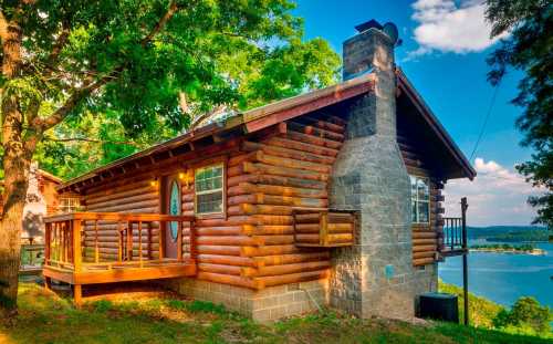 A cozy log cabin with a wooden deck, surrounded by trees and overlooking a serene lake under a blue sky.