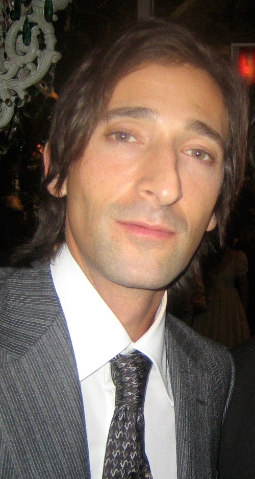 A man with long dark hair and a suit poses for the camera, looking directly with a neutral expression.