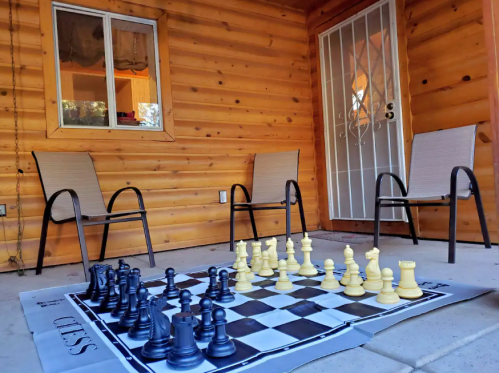 A large chessboard set up outside a wooden cabin, with black and yellow pieces and two chairs nearby.