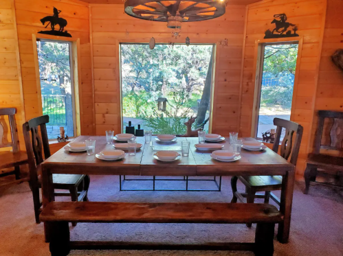 A rustic dining room with a wooden table set for a meal, surrounded by large windows showcasing a natural view.