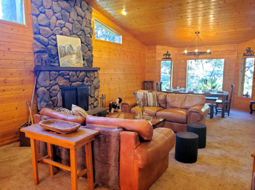 Cozy living room with stone fireplace, leather sofas, wooden accents, and dining area in a warm, rustic cabin setting.