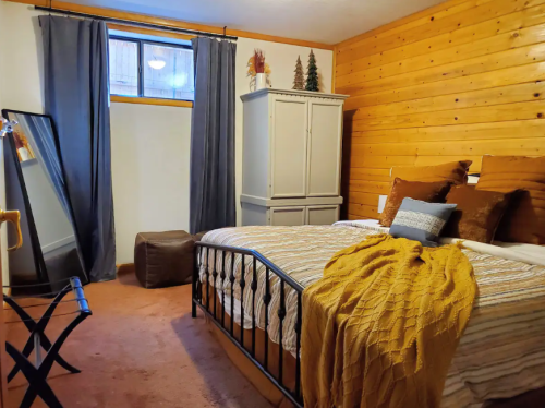 Cozy bedroom with wooden walls, a bed with a yellow throw, a mirror, and a window with curtains.