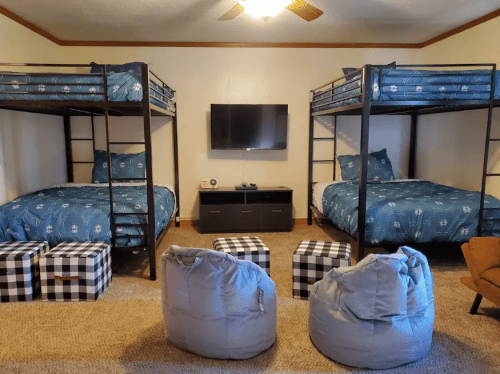 A cozy room with two bunk beds, a TV, and bean bags, featuring blue bedding and checkered ottomans.