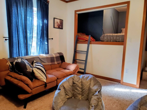 Cozy room featuring a sofa, bean bag chair, and a lofted bed accessed by a ladder, with blue curtains and warm lighting.