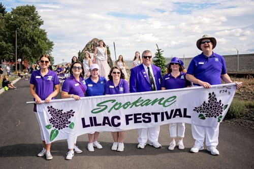 A group of people in purple shirts holding a banner for the Spokane Lilac Festival, smiling and posing for the photo.