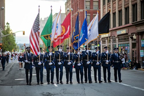 A color guard in uniform holds various flags, including the American flag, during a parade on a city street.