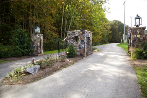 Stone entrance gates with lanterns, leading to a tree-lined road and landscaped garden.