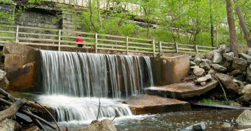 A small waterfall cascades over rocks, surrounded by greenery, with a person in red standing nearby on a wooden path.