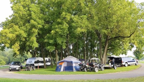A campsite with tents, RVs, and ATVs surrounded by lush green trees in a circular layout.