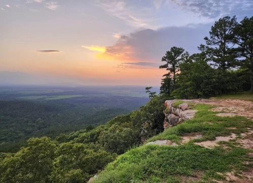 A scenic view from a rocky overlook, showcasing a sunset over lush green valleys and distant hills.