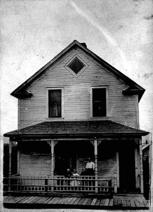 A vintage black-and-white photo of a two-story wooden house with a porch, featuring two people standing in front.