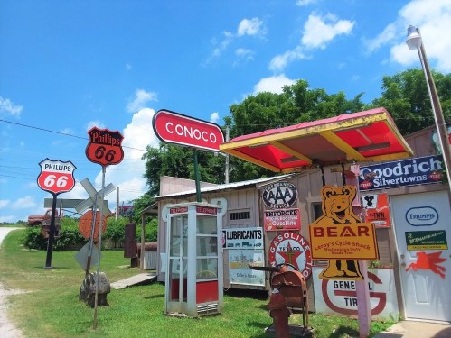 Vintage gas station signs, including Conoco and Phillips 66, with a rustic building and blue sky in the background.