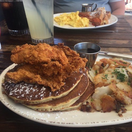 A plate of pancakes topped with fried chicken, served with hash browns and a small cup of syrup. Drinks in the background.
