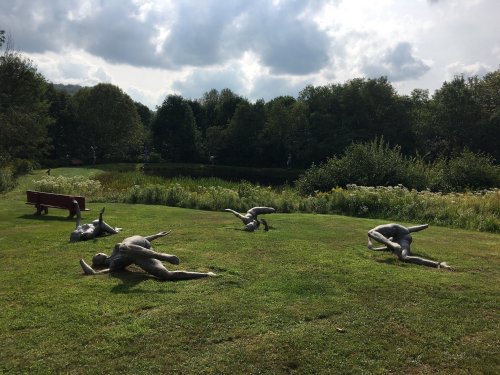 Sculptures of human figures lying on grass near a pond, surrounded by trees and a cloudy sky.