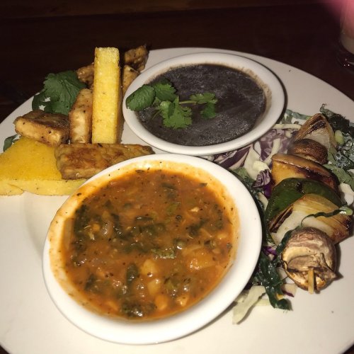 A plate featuring polenta, black bean sauce, vegetable stew, and a skewer of grilled vegetables, garnished with cilantro.