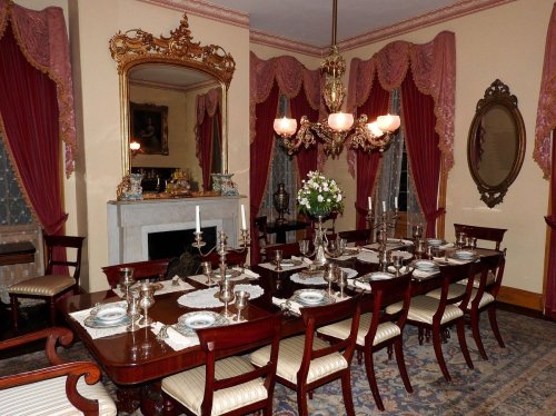 Elegant dining room with a long table set for a meal, ornate chandeliers, and rich drapery.