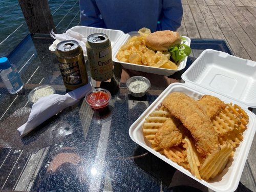 Two takeout containers with fried fish and waffle fries, a burger, chips, and drinks on a table by the water.