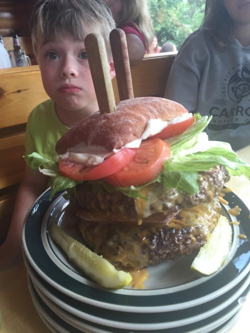 A large, stacked burger with lettuce, tomato, and pickles, next to a surprised child in a restaurant setting.