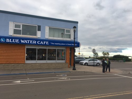 A blue and wooden café with a sign reading "Blue Water Cafe" and two people standing outside on a cloudy day.