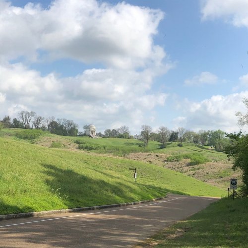A winding road leads through green hills under a partly cloudy sky, with a building visible in the distance.