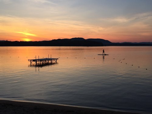 A serene sunset over a calm lake, with a silhouette of a person paddleboarding near a dock.