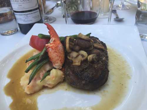 A gourmet plate featuring steak topped with mushrooms, lobster, green beans, and creamy mashed potatoes, with red wine in the background.