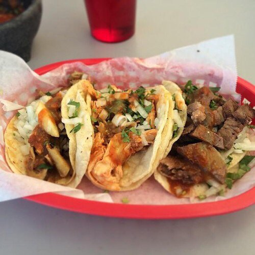 Three tacos in a red basket, filled with meat, onions, and cilantro, served with a side of salsa.
