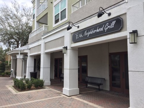 Exterior view of Teak Neighborhood Grill, featuring a sign, benches, and a brick pathway in a landscaped area.
