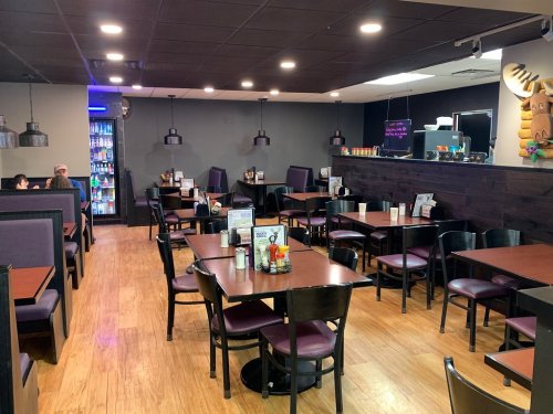 A spacious restaurant interior with wooden floors, tables, and chairs, featuring a drink fridge and a cozy atmosphere.