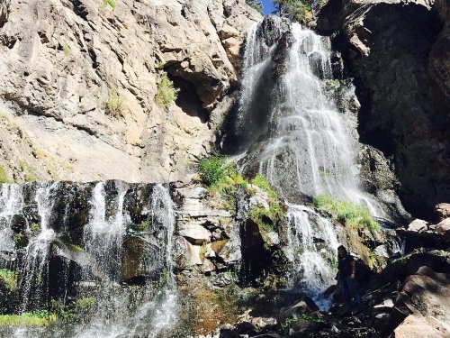 A cascading waterfall flows down rocky cliffs, surrounded by greenery and sunlight. A person stands near the base.