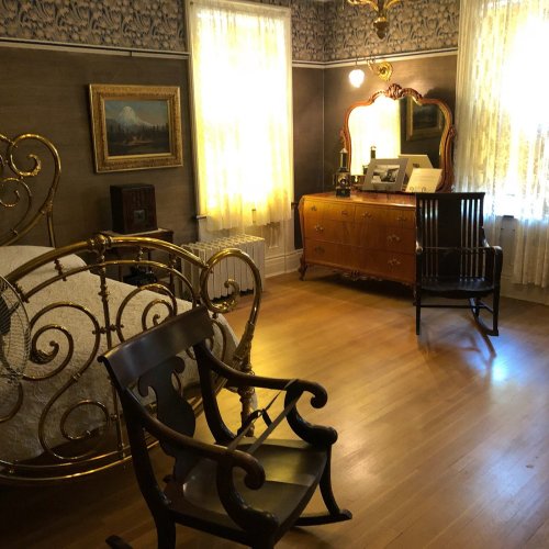 A vintage bedroom featuring a brass bed, wooden dresser, and two chairs, with soft light filtering through lace curtains.