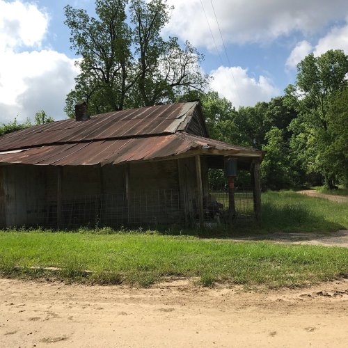 An old, weathered barn with a rusty roof, surrounded by lush greenery and a dirt road under a partly cloudy sky.