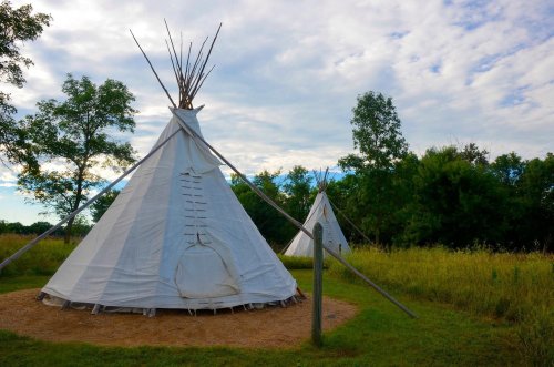 Two white tipis stand in a grassy area, surrounded by trees under a partly cloudy sky.