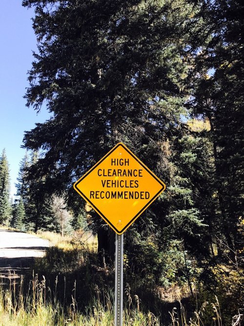 A yellow road sign reads "High Clearance Vehicles Recommended," surrounded by trees and a clear blue sky.