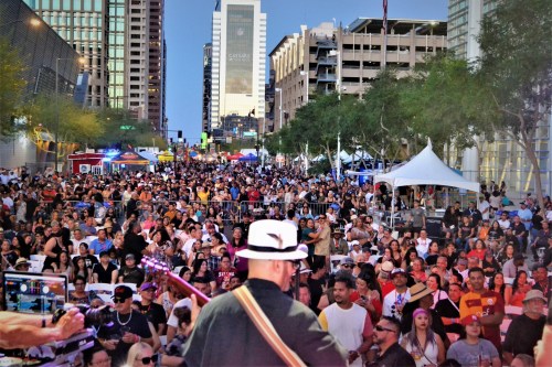 A large crowd enjoys a live music performance in an urban setting, with buildings and tents in the background.