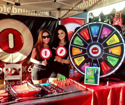 Two women in black shirts stand at a promotional booth, holding signs and smiling next to a colorful prize wheel.