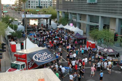A lively outdoor event with a crowd, food tents, and a stage, set on a city street surrounded by buildings.