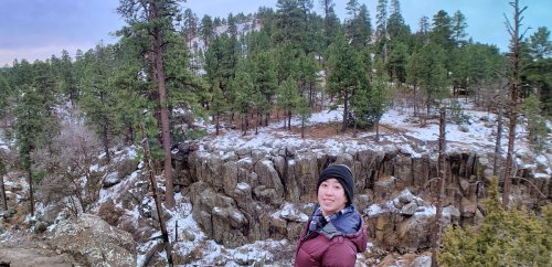 A person stands on rocky terrain surrounded by snow and pine trees, smiling at the camera in a scenic outdoor setting.