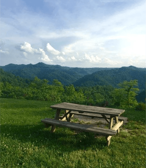 A wooden picnic table sits on green grass, overlooking rolling hills and a blue sky with fluffy clouds.