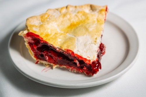 A slice of cherry pie on a white plate, featuring a flaky crust and vibrant red filling.