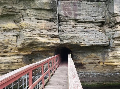A wooden bridge leads into a rocky tunnel surrounded by layered stone formations.
