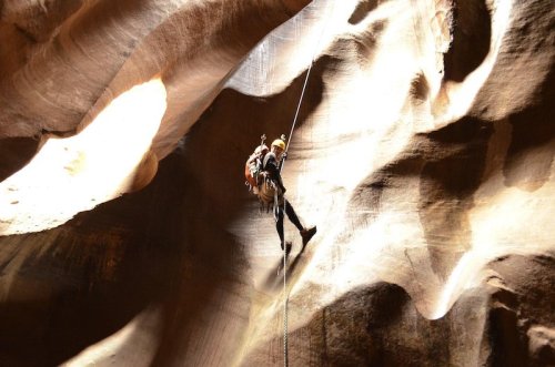 A climber rappels down a textured rock face in a narrow canyon, illuminated by natural light.