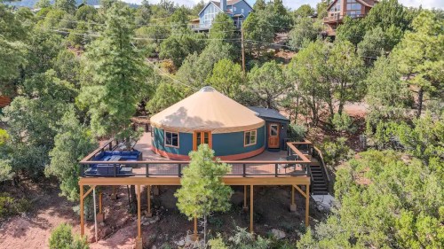 A colorful yurt on a raised deck surrounded by trees, with nearby houses in a scenic outdoor setting.