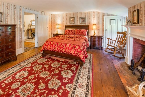 Cozy bedroom with a wooden bed, red floral bedding, rocking chair, and vintage decor against patterned wallpaper.