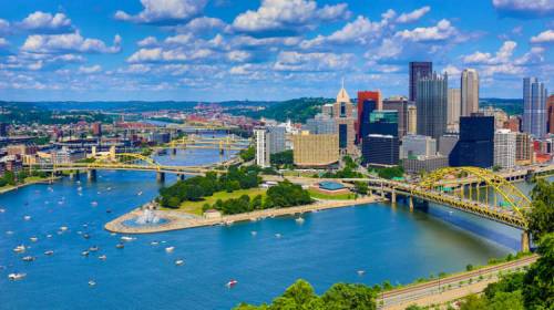 A vibrant cityscape of Pittsburgh, featuring rivers, bridges, and a skyline under a blue sky with fluffy clouds.