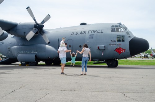 A family approaches a U.S. Air Force C-130 aircraft on a sunny day, with one child being carried on a parent's back.